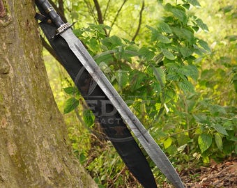 May include: A double-edged sword with a metallic blade featuring a central fuller, a black handle with silver-toned accents, and a black leather sheath. The sword is positioned diagonally against a tree trunk in a natural outdoor setting.