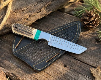 May include: A fixed-blade knife with a textured metal blade, a bright green spacer, and a brown antler handle. The knife rests on a black leather sheath featuring a repeating embossed pattern and yellow stitching along the outer edges.
