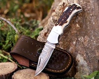 May include: A folding pocket knife with a patterned Damascus steel blade and a textured brown antler handle. The knife is positioned next to a dark brown leather sheath featuring decorative embossed patterns and a metal snap button closure.