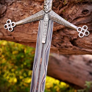 May include: A sword featuring a dark, layered Damascus steel blade with visible wavy patterns. The metal crossguard displays intricate scrollwork, a central lion emblem, and four-ringed terminals at each end of the quillons.