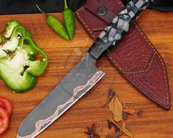 May include: A kitchen knife with a dark grey blade featuring a wavy copper-colored inlay and a handle made of black and white mosaic pieces. The knife is accompanied by a brown leather sheath with decorative stitching and a snap button closure.