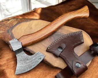 May include: A hand axe with a curved wooden handle and a patterned Damascus steel blade. The axe is accompanied by a dark brown leather sheath featuring decorative stitching and two metal snap buttons for secure closure and storage.