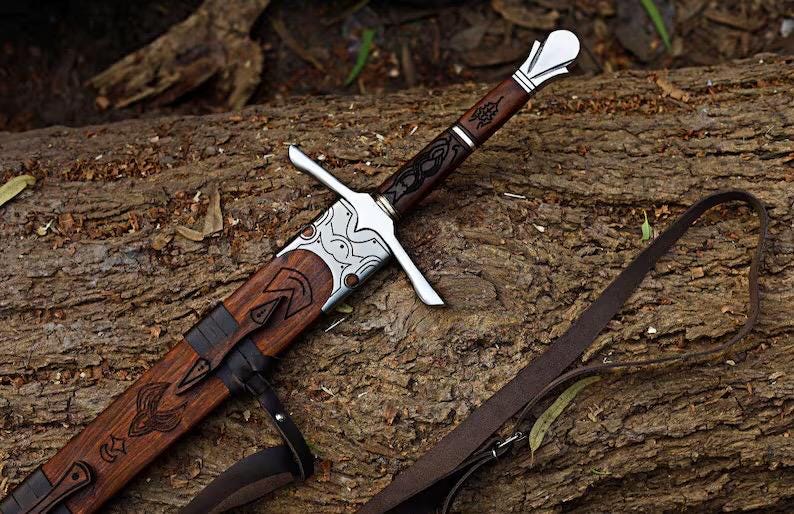 May include: A sword with a dark brown wooden handle and scabbard, featuring silver-toned metal accents on the pommel, crossguard, and throat. The scabbard displays intricate dark engravings and is attached to a long, thin brown leather strap.