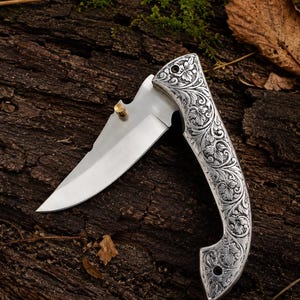 May include: A folding pocket knife with a silver-toned handle featuring intricate floral scrollwork engravings. The blade is made of polished steel and includes a small, cylindrical brass-coloured thumb stud for opening the mechanism.