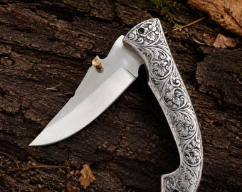 May include: A folding pocket knife with a silver-toned handle featuring intricate floral scrollwork engravings. The blade is made of polished steel and includes a small, cylindrical brass-colored thumb stud for opening the mechanism.