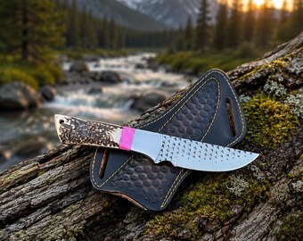 May include: A fixed-blade knife with a textured metal blade, a bright pink band at the bolster, and a dark brown antler handle. The knife rests on a black leather sheath featuring a honeycomb pattern and visible yellow stitching along the edges.