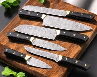 May include: A set of six kitchen knives with patterned steel blades and black handles. The knives are arranged on a wooden cutting board, with gold-colored bolsters and rivets. The blades have a unique, textured pattern.
