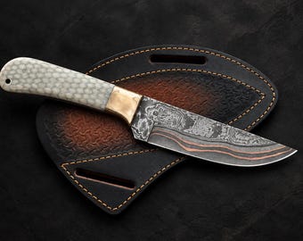 May include: A fixed-blade knife featuring a patterned Damascus steel blade with visible copper layers, a brass bolster, and a textured off-white handle. The knife rests on a black and brown leather sheath with yellow stitching and two belt slots.