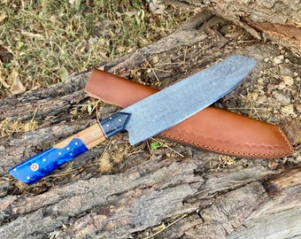 May include: A Damascus steel chef's knife with a blue and wood handle, resting on a wooden surface. The knife has a patterned blade and is accompanied by a brown leather sheath. The handle features a blue resin design with white dots and a red emblem.