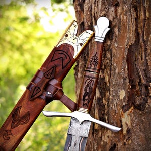 May include: A dagger with a patterned steel blade, a wooden handle featuring intricate carvings, and a polished silver pommel. The matching wooden scabbard displays dark wood grain, engraved symbols, and a decorative gold-toned metal throat.