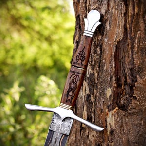 May include: A dagger with a dark wood handle featuring intricate black etched patterns, a polished silver-toned pommel, a matching silver-toned crossguard, and a patterned metal blade with a dark, textured finish along the central section.