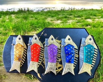 May include: A decorative wall hanging featuring five colorful fish sculptures. Each fish has a unique pattern and color, including gold, red, purple, blue, and teal. The fish are mounted on a black wooden plaque with a scenic background.