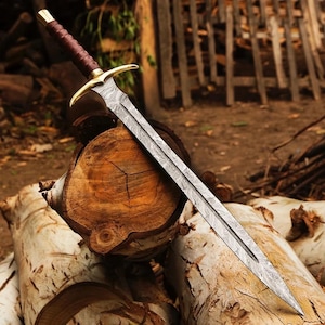 May include: A double-edged sword featuring a patterned Damascus steel blade, a gold-toned metal crossguard, and a dark brown leather-wrapped handle with a gold-toned pommel cap. The blade displays a distinct wavy, layered metal texture.