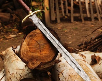 May include: A double-edged sword featuring a patterned Damascus steel blade, a gold-toned metal crossguard, and a dark brown leather-wrapped handle with a gold-toned pommel cap. The blade displays a distinct wavy, layered metal texture.