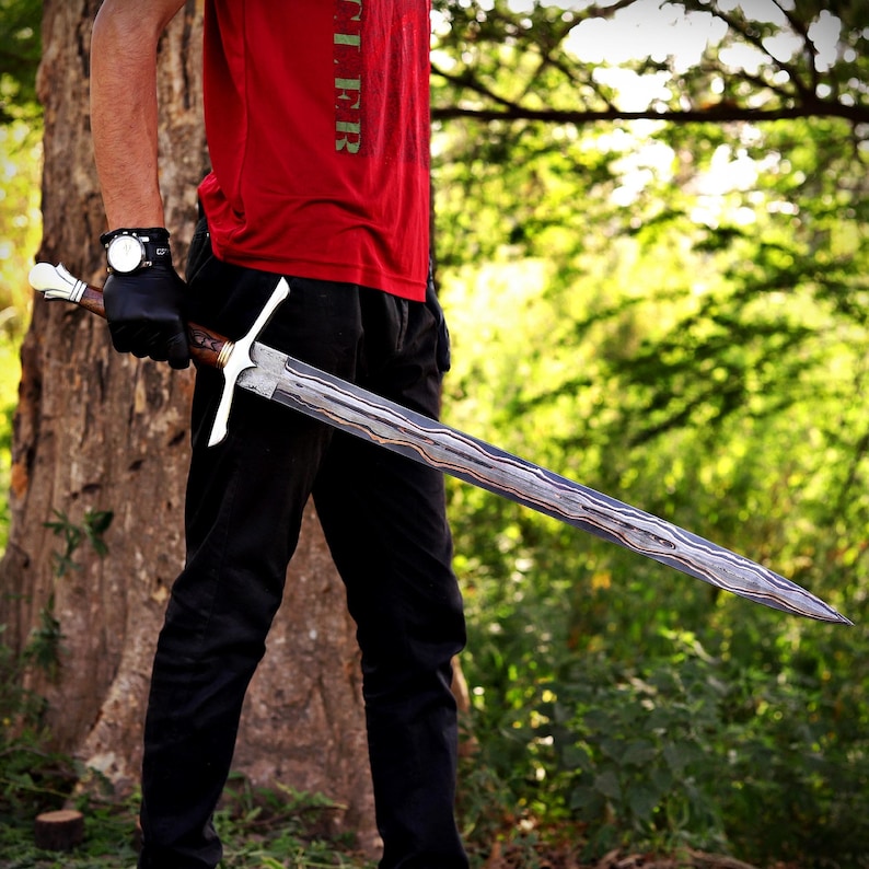 May include: A long sword featuring a double-edged blade with a distinct wavy, layered Damascus steel pattern. The hilt includes a silver-toned crossguard, a dark brown wooden grip with gold-colored accents, and a white, rounded pommel at the end.