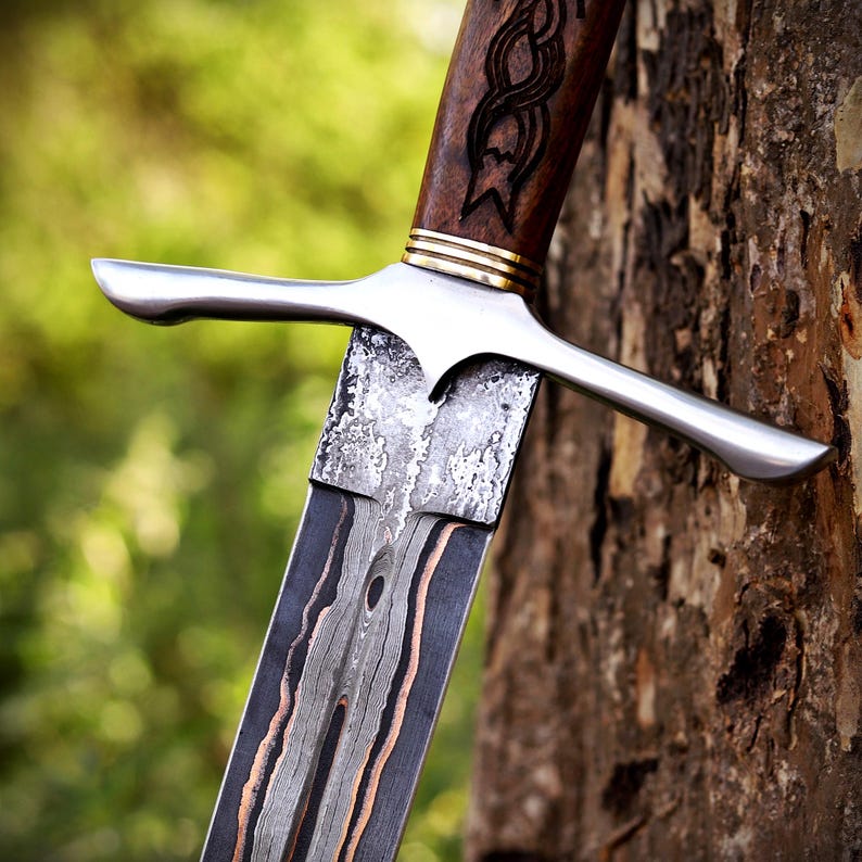 May include: A sword with a patterned Damascus steel blade featuring dark and copper-colored layers. The hilt has a polished silver-colored crossguard, a gold-colored ring, and a dark brown wooden handle with an intricate carved knot design.