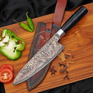 May include: A chef knife with a patterned Damascus steel blade and a dark, polished handle, resting on a brown leather sheath. The knife is accompanied by two green chilli peppers, sliced green bell pepper pieces, and tomato slices.