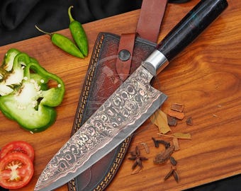May include: A chef knife with a patterned Damascus steel blade and a dark, polished handle, resting on a brown leather sheath. The knife is accompanied by two green chili peppers, sliced green bell pepper pieces, and tomato slices.