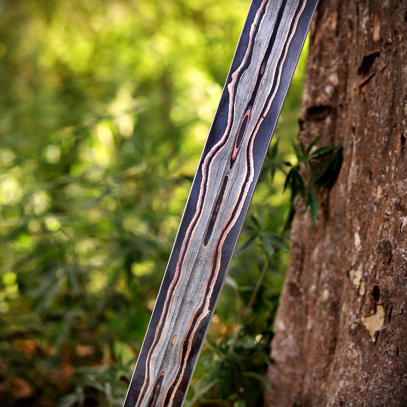 May include: A long, narrow metal blade featuring a distinct layered pattern of dark charcoal, light silver, and copper-toned bands running vertically along its length. The surface displays a complex, organic, flowing texture of contrasting metal.