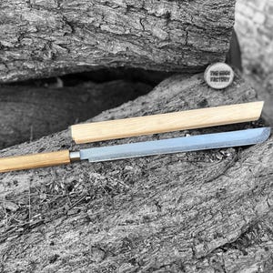 May include: A Japanese-style knife with a wooden handle and a silver blade, resting on a textured rock surface. The knife is accompanied by a wooden sheath. A small, round wooden emblem with the text "THE EDGE FACTORY" is also present.