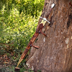 May include: A sword with a silver-toned crossguard and pommel, featuring a brown leather scabbard adorned with intricate dark engravings, brass-toned metal accents at the tip and throat, and leather straps for attachment, resting against a tree.