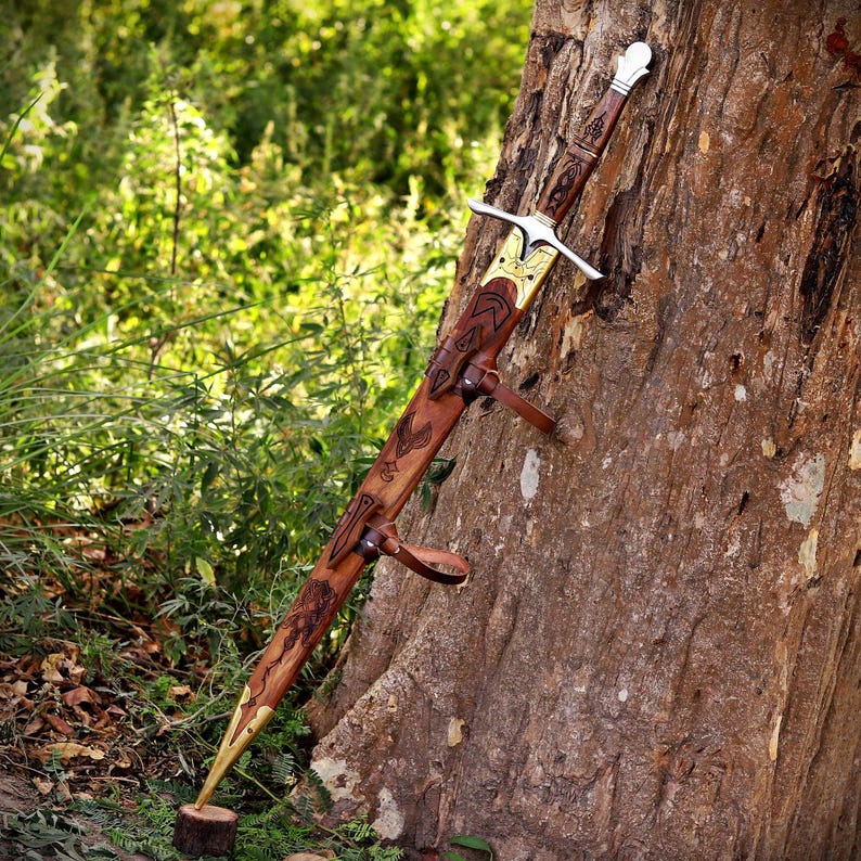 May include: A sword with a silver-toned crossguard and pommel, featuring a brown leather scabbard adorned with intricate dark engravings, brass-toned metal accents at the tip and throat, and leather straps for attachment, resting against a tree.