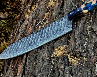 May include: A large, ornate chef's knife with a patterned blade and a decorative handle. The blade has a textured, wave-like design. The handle features blue, gold, and red accents. The knife rests on a textured, brown tree trunk.