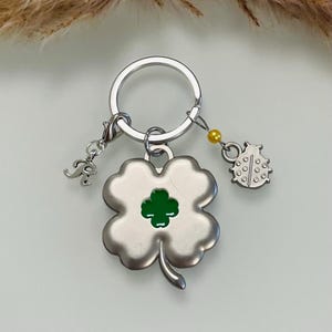 Puede incluir: Llavero plateado con un dije de trébol de cuatro hojas con centro verde, un dije de mariquita y un pequeño dije inicial. El llavero tiene un anillo plateado y un acento de cuentas amarillas. El fondo es de color claro y neutro.