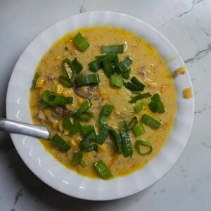 May include: A white bowl filled with a creamy yellow corn chowder, topped with chopped green onions. The soup contains visible corn kernels and small pieces of meat. A silver metal spoon rests inside the bowl, which is set against a white surface.