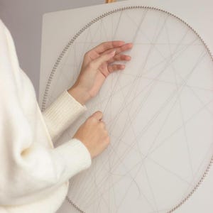 Può includere: Una persona che lavora a un progetto di string art. L'opera presenta uno sfondo bianco con un semicerchio di piccoli chiodi argentati. Un filo grigio sottile è teso sulla superficie, creando un disegno geometrico.