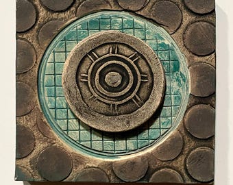 May include: Square ceramic tile with a textured surface. The tile features a central circular design with concentric circles and radial lines, surrounded by a turquoise square grid and brown circles. The tile has a rustic, earthy aesthetic.