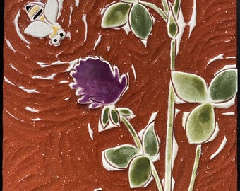 May include: Square ceramic tile with a bee, purple flower, and green leaves on a reddish-brown background. The bee is white with yellow and black details. The flower is a deep purple, and the leaves are green.
