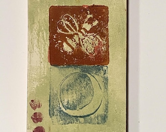 May include: Rectangular art piece with a light green background and various stamped designs. A yellow sun is at the top, a brown square with a white floral design in the center, and a blue crescent moon at the bottom. Three maroon dots are on the left.