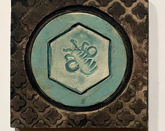 May include: Square ceramic tile with a teal hexagon in the center, featuring a bee design. The hexagon is surrounded by a teal circle and a dark brown square frame with a repeating quatrefoil pattern. The tile has a rustic, handmade appearance.