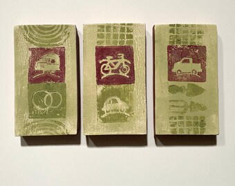 May include: Three rectangular, light green decorative tiles with red and green designs. One tile features a camper and wedding rings, another a bicycle and car, and the last a truck and gardening tools.