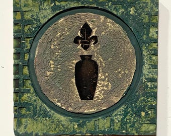 May include: Square, textured artwork with a green, grid-like border. A central circle features a dark vase and fleur-de-lis design. The artwork has a rustic, aged appearance, with a mix of green, brown, and black tones. The piece is likely a decorative item.