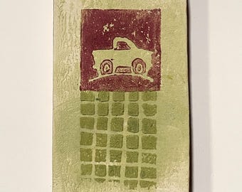 May include: Rectangular art piece with a light green background. A maroon square features a car silhouette. Below, a grid of green squares adds texture. The piece has a rustic, handmade appearance.