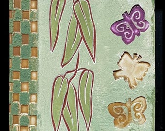 May include: Square ceramic tile with a light green background. The tile features a woven pattern on the left side in shades of green and brown. The center has a design of green leaves with red outlines, and three embossed butterflies in purple, tan, and gold.