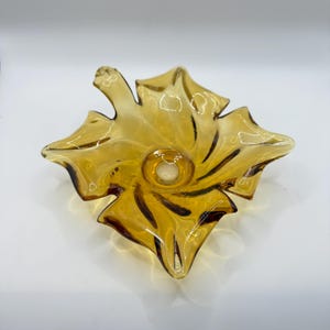 May include: A decorative amber-colored glass bowl in a leaf shape. The bowl has a central, circular indentation and dark brown accents along the edges of the leaf-shaped points. The glass is transparent, allowing light to pass through.