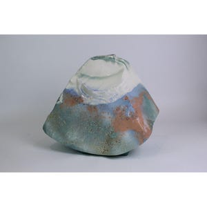 May include: A sculptural ceramic vase with an irregular, organic shape. The surface features a blend of white, light blue, and earthy brown glazes with a textured, speckled finish. The top section is primarily white, transitioning into blue and brown tones.
