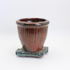 May include: A ceramic planter with a reddish-brown exterior and a dark brown rim. The planter has vertical grooves and sits on a square, turquoise-colored base. The pot is designed for indoor or outdoor use.