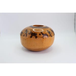May include: A round, tan ceramic vase with a dark brown rim and a central opening. The vase is decorated with dark brown figures and animal shapes around the body. The vase has a smooth, glossy finish.