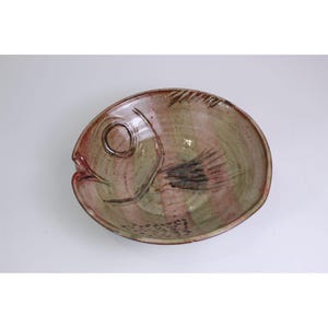 May include: A round ceramic bowl featuring a stylized fish design etched into the surface. The bowl has a mottled glaze in shades of muted green, brown, and pink, with a dark brown outline defining the fish eye, fins, and scales on the interior.