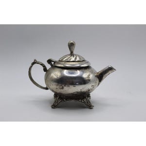 May include: A small, antique silver teapot with a rounded body, a curved handle, and a spout. The lid is topped with a decorative finial. The teapot rests on three ornate feet. A classic piece for tea lovers.