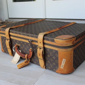 May include: A rectangular brown suitcase featuring a repeating monogram pattern in a lighter brown tone. The luggage includes tan leather corner reinforcements, two vertical leather buckle straps, a top handle, and a small attached luggage tag.