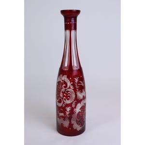 May include: A tall, slender glass decanter featuring a deep ruby red color. The neck is clear with vertical fluted cuts, while the base displays intricate, etched white floral and scrollwork patterns that contrast against the vibrant red glass.