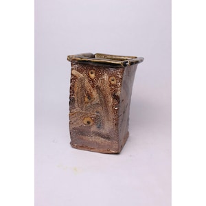 May include: A rectangular ceramic vase with a textured, brown surface. The vase features a dark brown rim and decorative patterns with circular accents. The design includes vertical and horizontal brushstrokes, creating a rustic aesthetic. The vase is suitable for home decor.