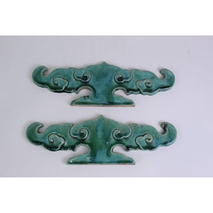 May include: Two matching decorative ceramic ornaments in a deep teal green glaze. Each piece features a stylized cloud-like silhouette with symmetrical, swirling scrollwork patterns and a flat base, showcasing a glossy, reflective finish.