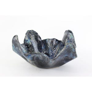 May include: A handcrafted, ceramic bowl with a unique, organic shape. The bowl features a dark, textured surface with iridescent blue and black hues. The rim has a wavy design, adding to its artistic appeal. The bowl is set against a white background.