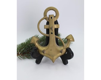 Può includere: Un'ancora in ottone con dettagli di corda, montata su un supporto in legno nero. L'ancora presenta un design classico con un anello in alto e due marre. Un rametto di sempreverde è sullo sfondo.